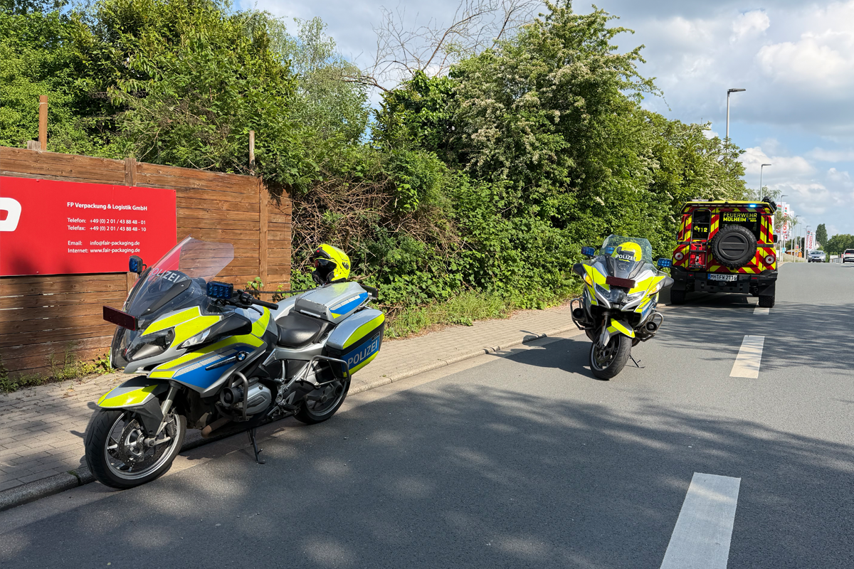 Zwei Polizeimotorräder stehen nebeneinander auf einer Straße. Ein Feuerwehrfahrzeug befindet sich weiter hinten auf der Straße. Es ist ein sonniger Tag.