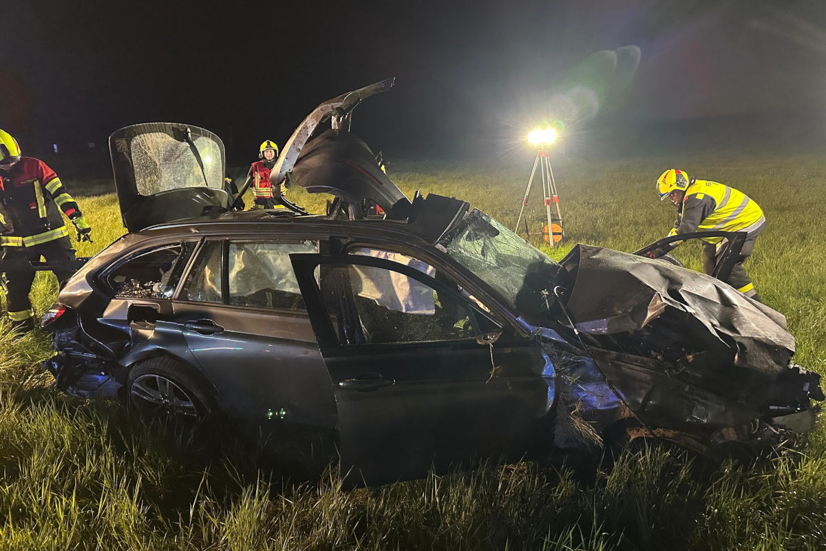 Ein stark beschädigtes Auto steht auf einer Wiese. Rettungskräfte in Schutzkleidung arbeiten daran, beleuchtet von einem Flutlicht.