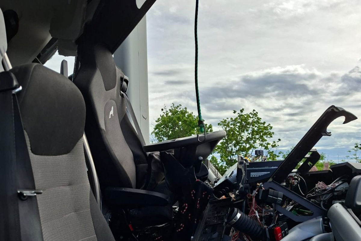 Das Bild zeigt die Innenansicht eines beschädigten Fahrzeugs. Die Elektronik liegt offen, und Teile des Cockpits sind zerstört. Im Hintergrund sind Bäume und ein bewölkter Himmel sichtbar.