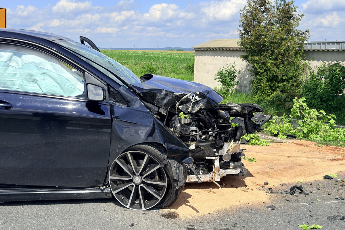 Ein stark beschädigtes schwarzes Auto steht am Straßenrand mit ausgeklapptem Airbag und zerstörter Front. Im Hintergrund sind Wiesen und ein Baum zu sehen.