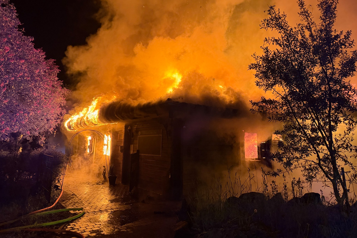 Ein Haus brennt in der Nacht intensiv, mit hellen Flammen und starkem Rauch. Die Umgebung ist dunkel, Bäume sind im Vordergrund sichtbar.