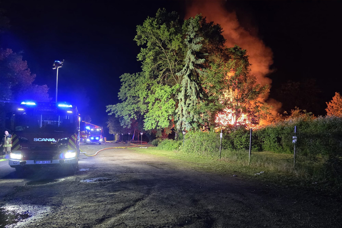 Zwei Feuerwehrfahrzeuge stehen nachts auf einer Straße. Im Hintergrund ist ein stark brennendes Waldstück zu sehen, das von Flammen erleuchtet wird.