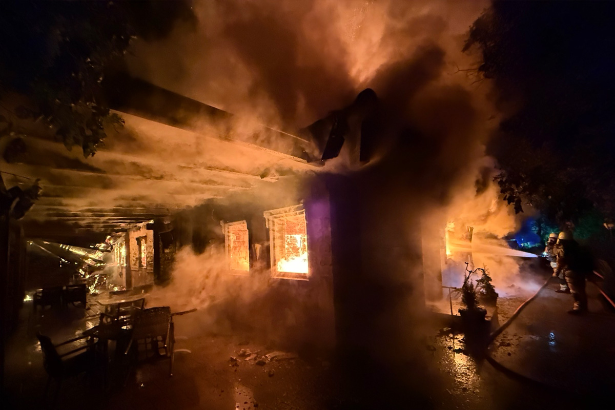 Nachts brennt ein Haus, aus dem dichter Rauch aus den Fenstern tritt. Feuerwehrleute sind mit Löscharbeiten beschäftigt.