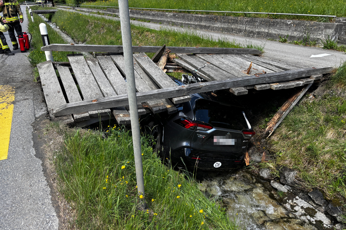 Ein schwarzes Auto ist unter einer eingestürzten Holzbrücke in einem Graben zu sehen. Ein Feuerwehrmann steht mit Feuerlöscher daneben.