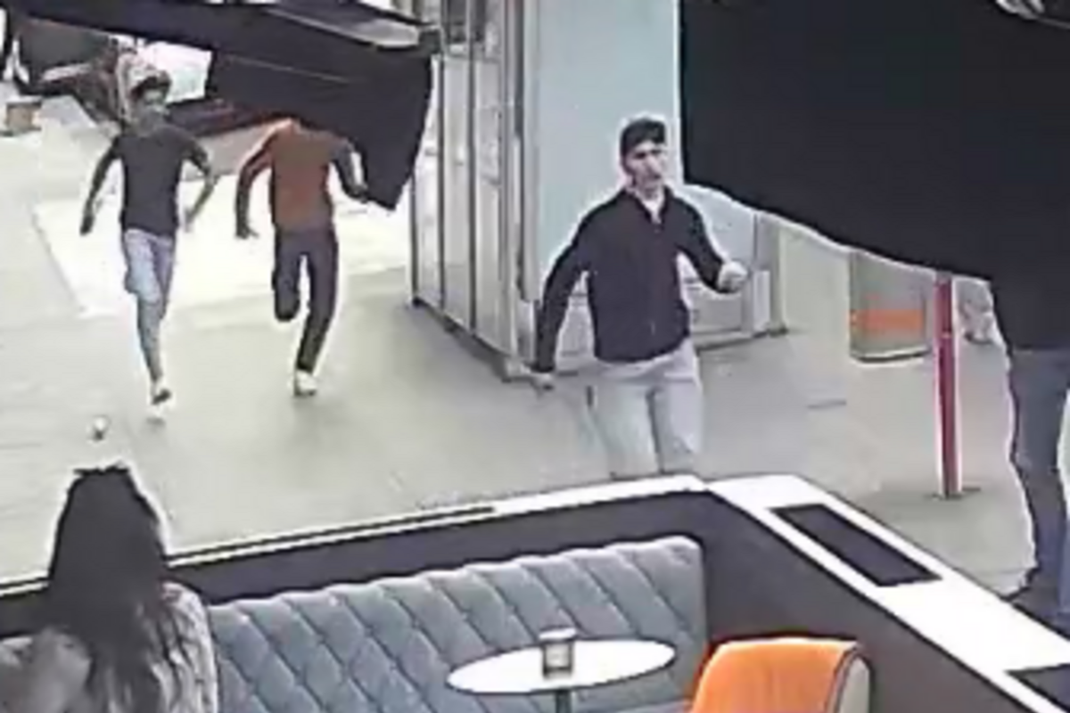 Drei junge Männer rennen auf einem Gehweg, während eine Frau auf einer Sitzbank im Vordergrund sitzt. Eine moderne Umgebung ist zu erkennen.