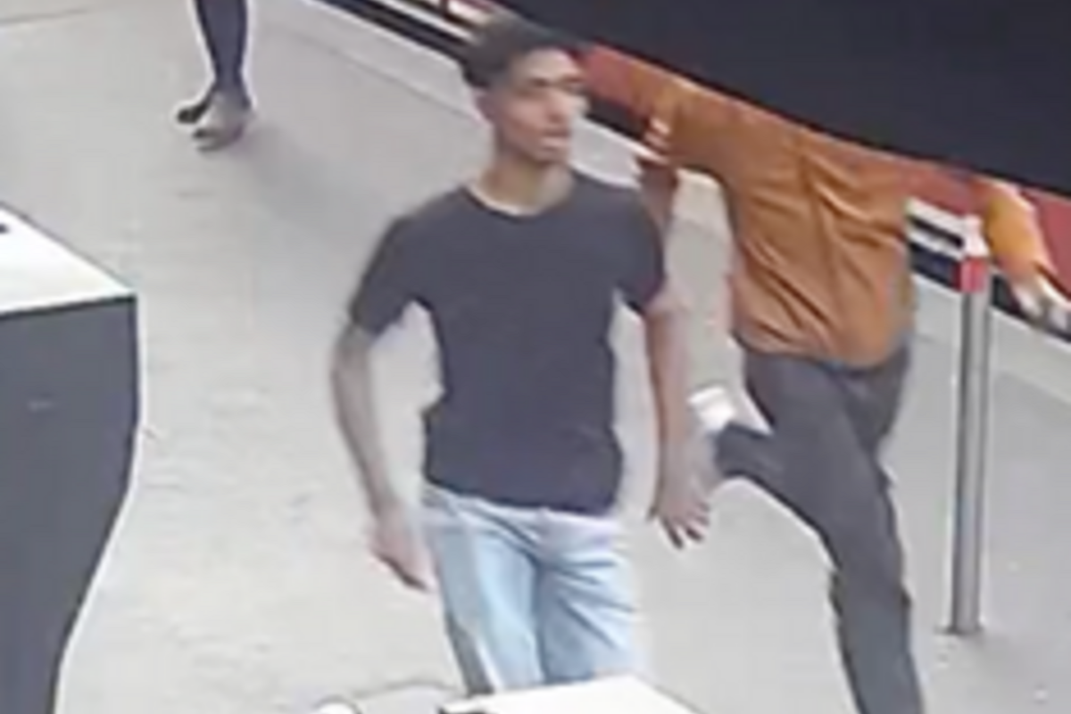 Zwei Männer sind in einem öffentlichen Innenbereich sichtbar. Eine Person trägt ein schwarzes T-Shirt und eine helle Jeans, die andere ist in einem orangefarbenen Oberteil gekleidet.