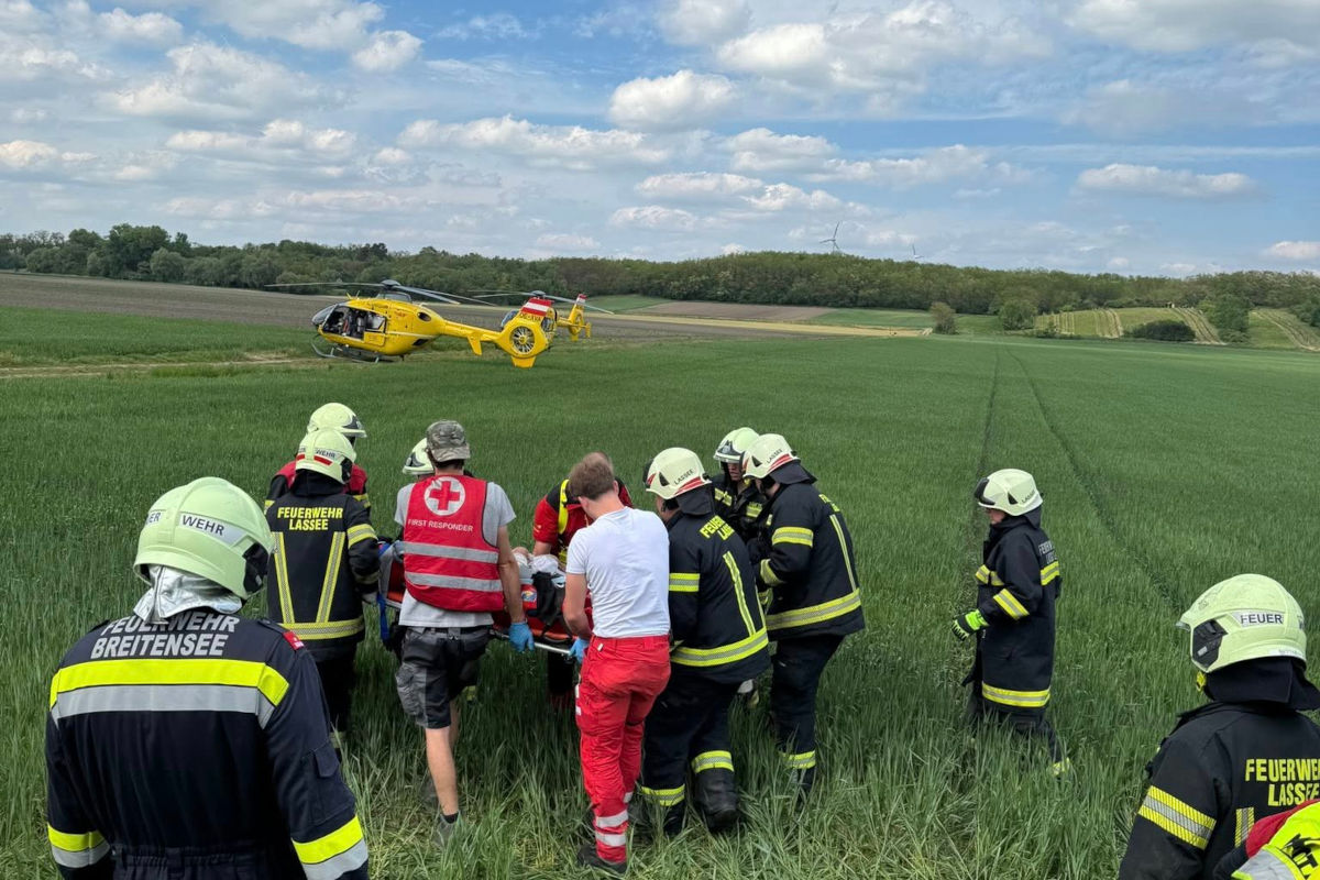 Auf einem Feld führen Rettungskräfte einen Einsatz durch. Ein gelber Hubschrauber ist im Hintergrund zu sehen und mehrere Personen tragen eine Trage.