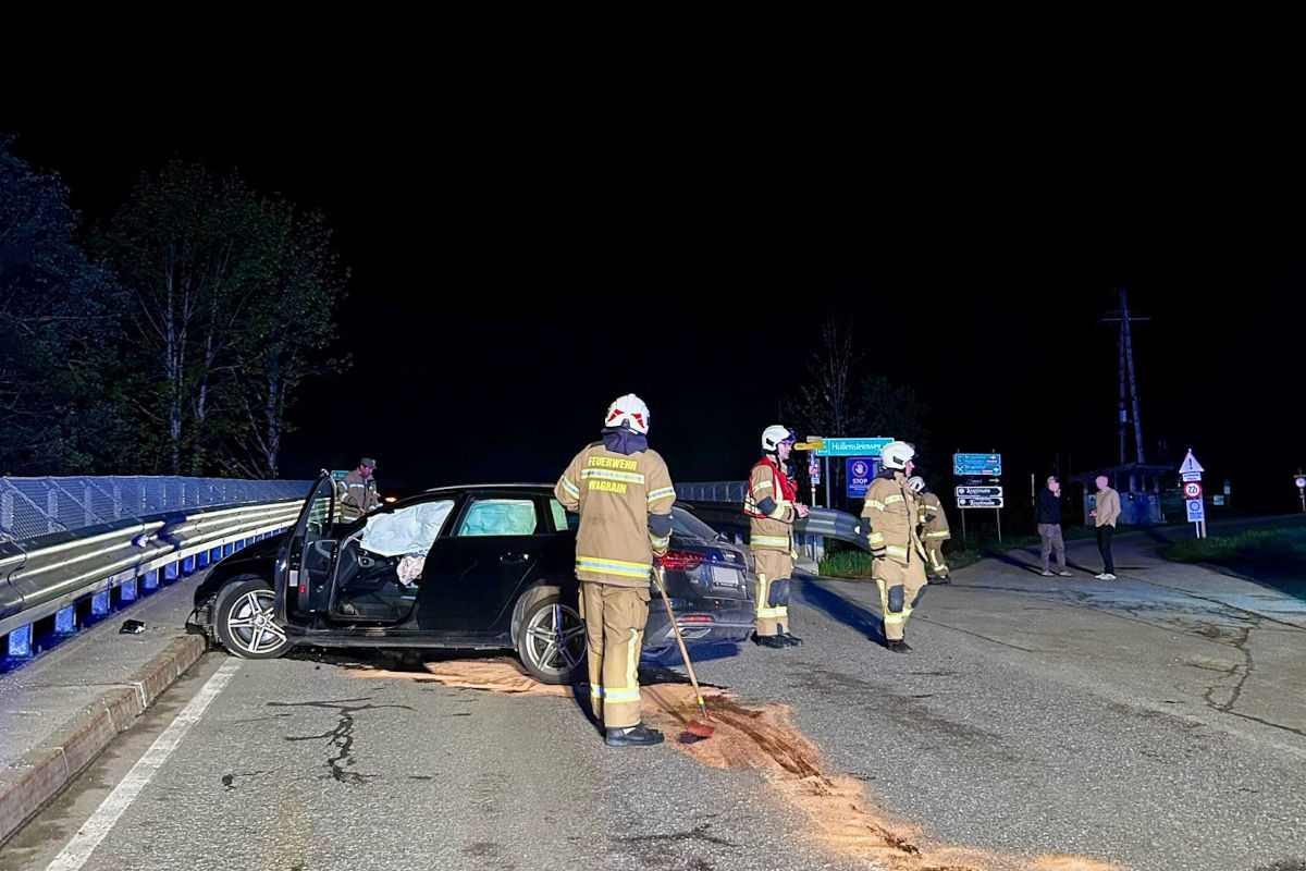 Ein schwarzes Auto mit beschädigter Vorderseite steht auf einer Straße. Feuerwehrleute in Uniform sind vor Ort und führen Aufräumarbeiten durch. Die Szene ist nachts beleuchtet.