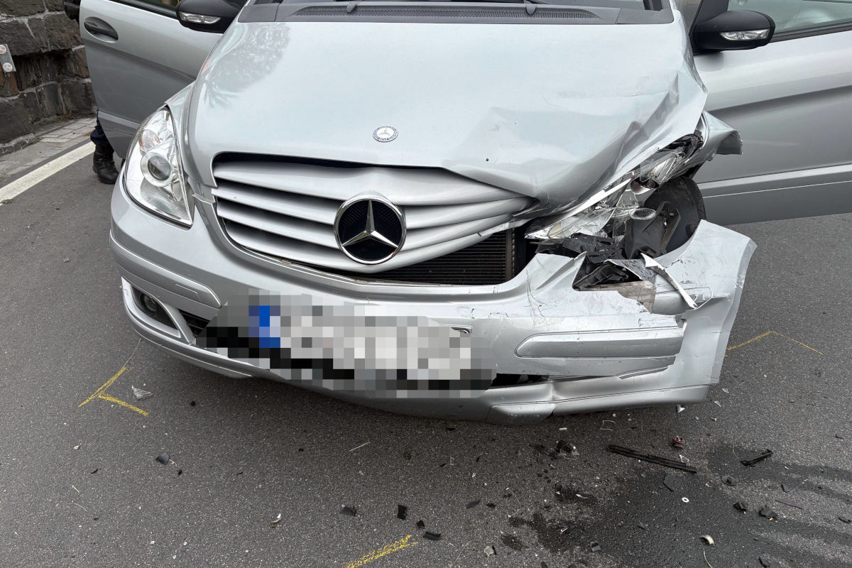 Ein silberner Mercedes, dessen rechte Vorderseite bei einem Unfall stark beschädigt wurde, steht auf einer Straße. Der Scheinwerfer ist zerbrochen und die Stoßstange ist eingedrückt.
