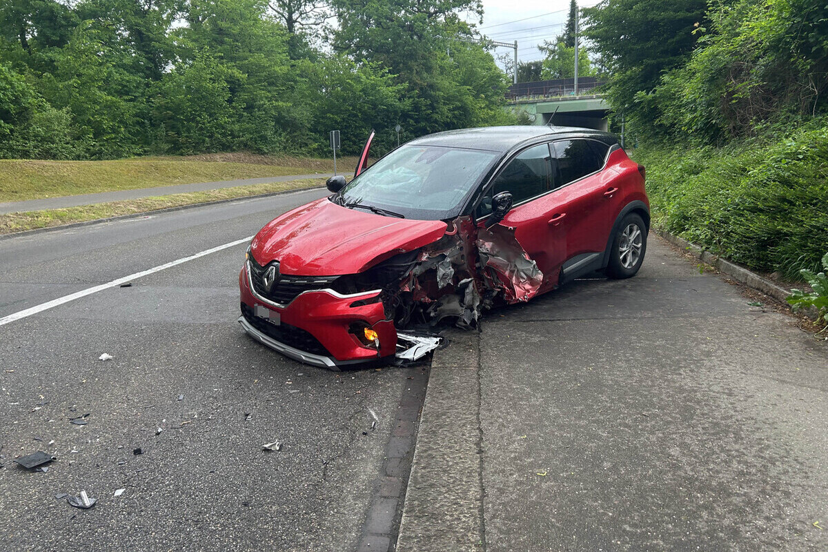 Ein rotes Auto steht mit starkem Frontschaden auf einem Gehweg neben einer Straße. Die Umgebung ist grün und bewaldet.