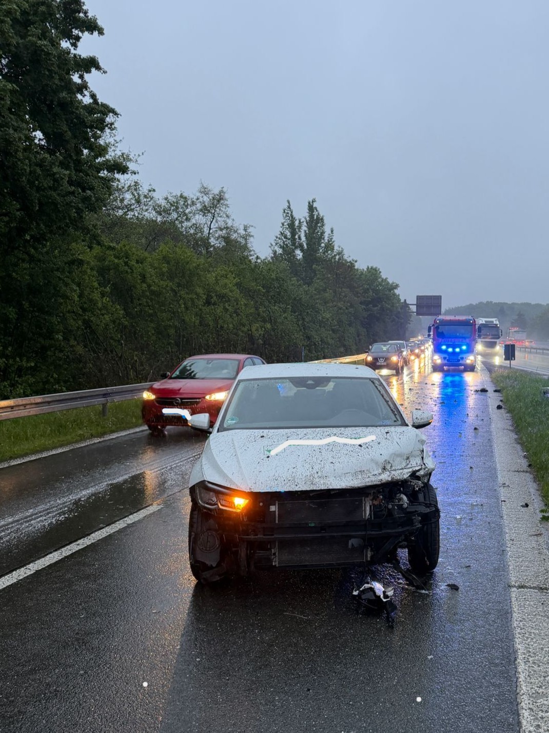 Ein beschädigtes Auto steht auf einer nassen Autobahn im Regen. Im Hintergrund sind weitere Fahrzeuge und Blaulicht zu sehen.