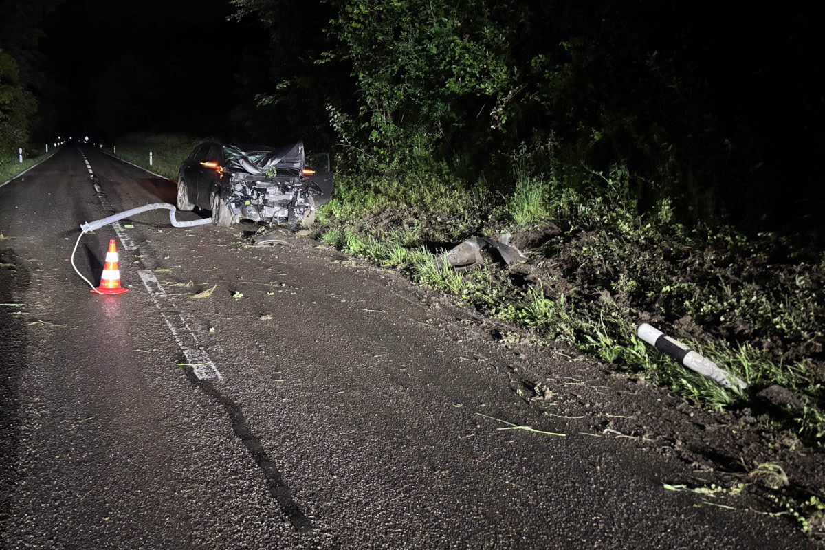 Ein stark beschädigtes Auto steht bei Nacht auf einer Landstraße. Ein Warnkegel und Trümmer sind sichtbar.