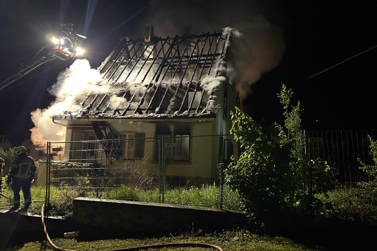 Bei Nacht löscht die Feuerwehr ein brennendes Haus. Rauch steigt vom Dach auf, während Feuerwehrleute arbeiten.