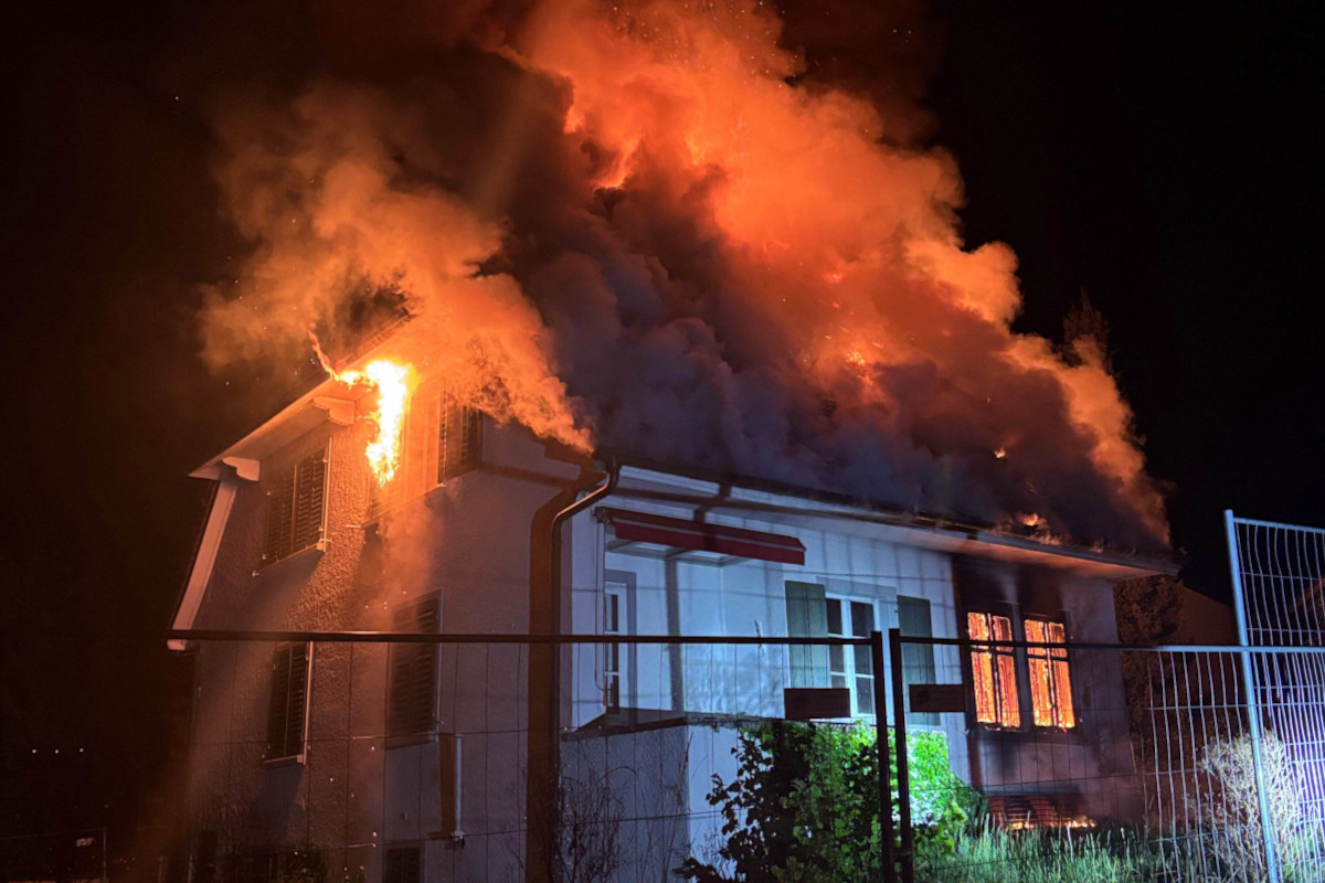 Ein zweistöckiges Haus brennt stark, Flammen und dichter Rauch steigen vom Dach auf. Es ist Nacht und das Szenario wird von einem bläulichen Licht beleuchtet.