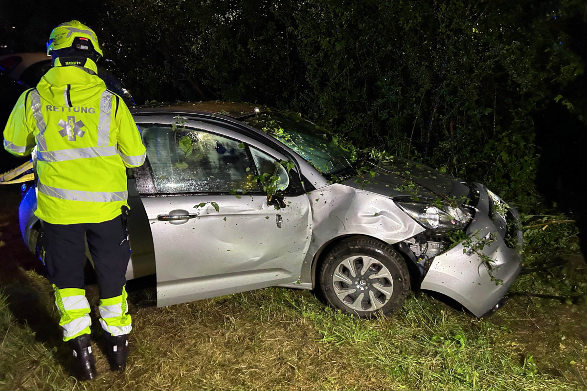 Ein graues Auto mit grünen Blättern darauf steht verbeult auf einer Wiese. Ein Rettungspersonal in gelber Uniform ist zu sehen.