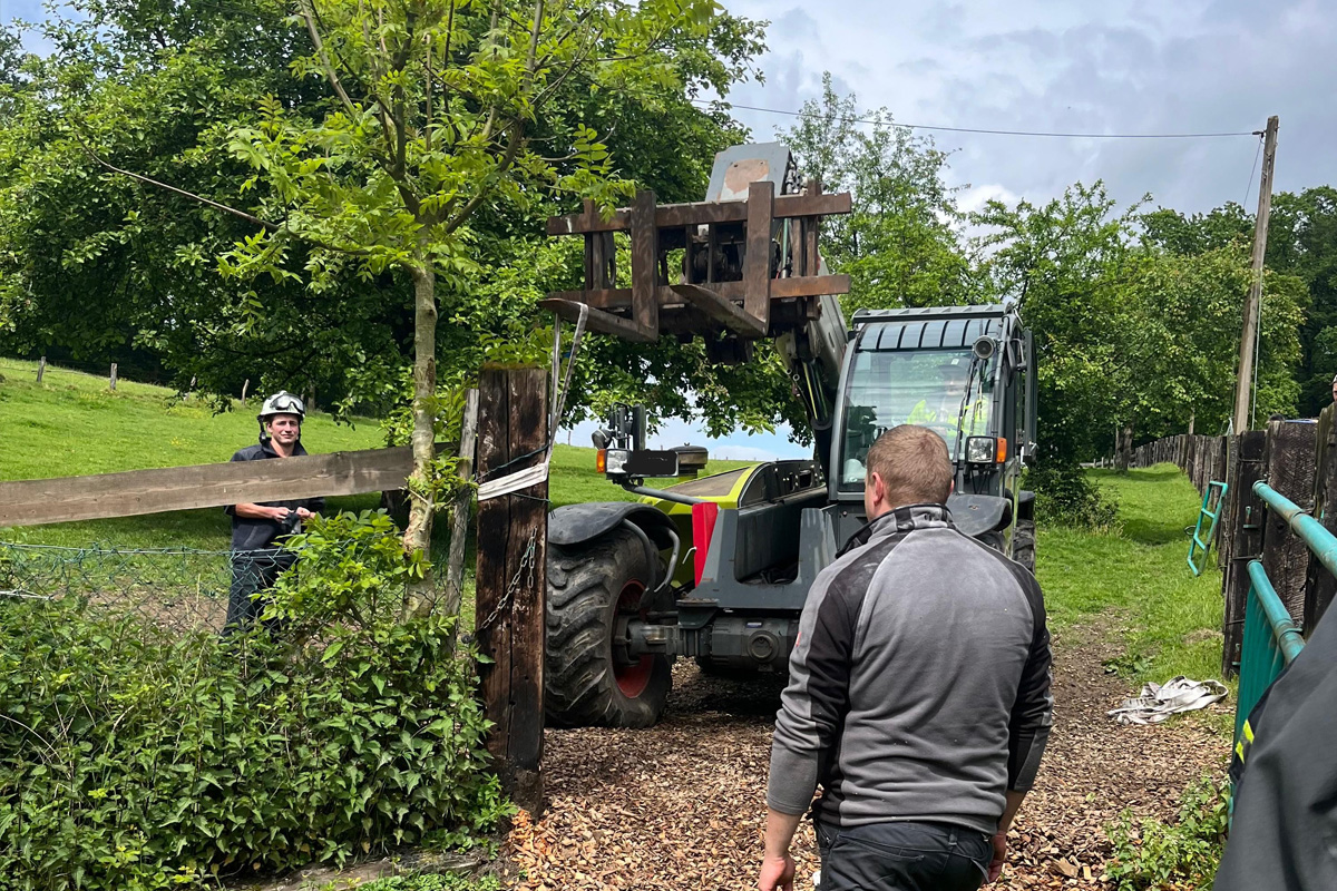 Ein Mann steht in einer ländlichen Umgebung und beobachtet einen Traktor mit einem großen Holzlader, der eine Arbeit ausführt. Im Hintergrund sind Bäume und ein Zaun zu sehen.