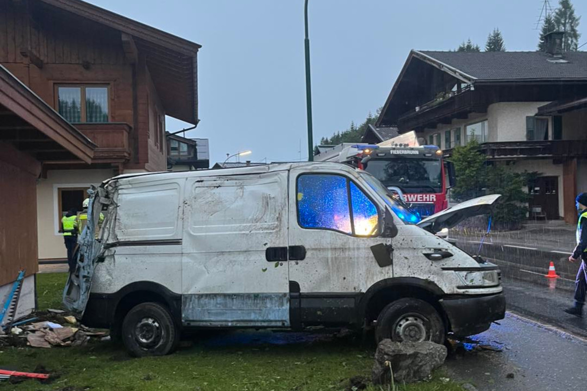 Ein beschädigter weißer Van steht auf einer Wiese vor einem Holzgebäude. Im Hintergrund ist ein Feuerwehrfahrzeug sichtbar.