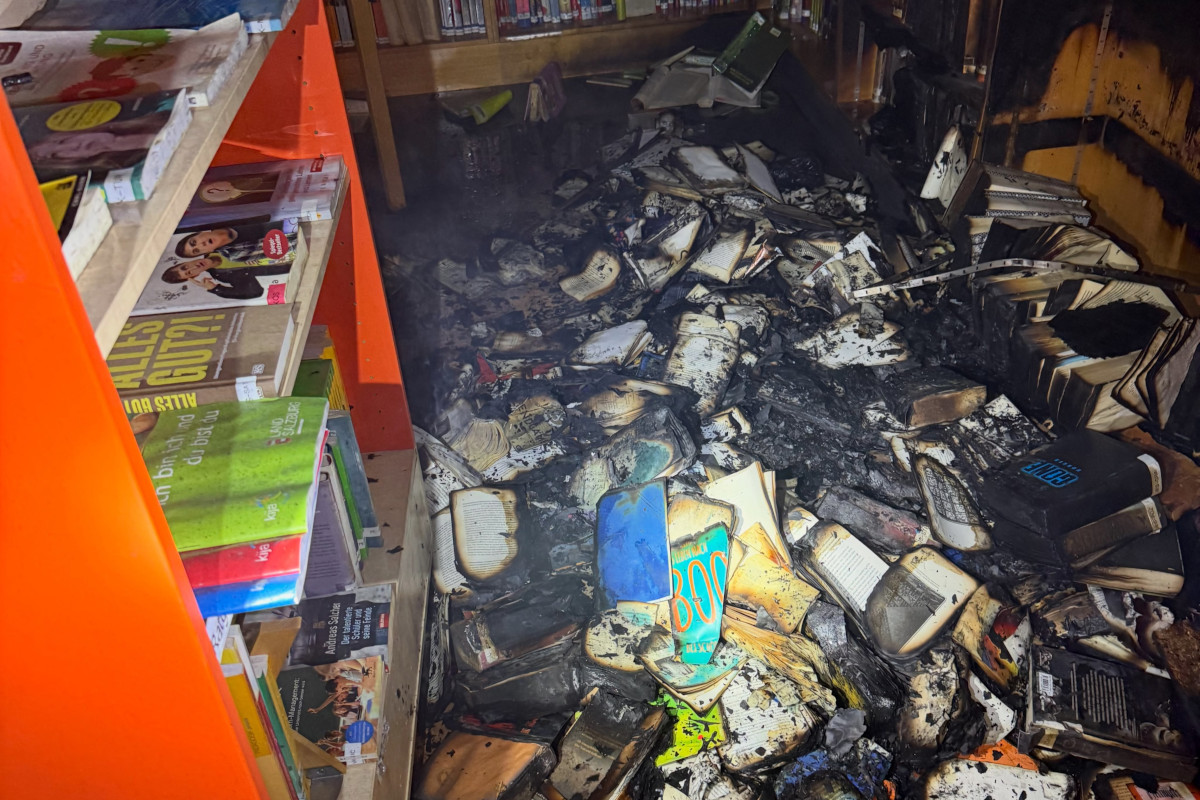 Ein Haufen verkohlter Bücher liegt auf dem Boden einer Bibliothek. Regale mit intakten Büchern sind im Hintergrund zu sehen.