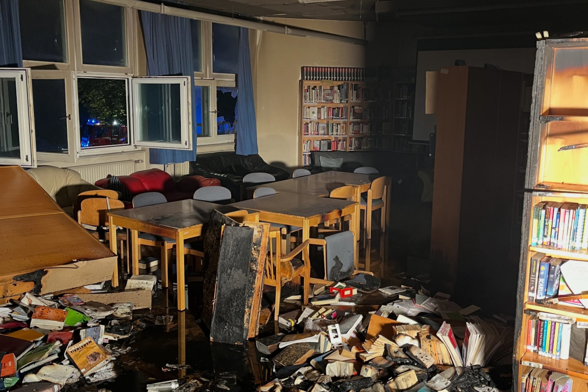 In einem Raum liegen verbrannte Bücher auf dem Boden, umgeben von Tischen und Stühlen. Am Fenster sind blaue Lichter sichtbar.