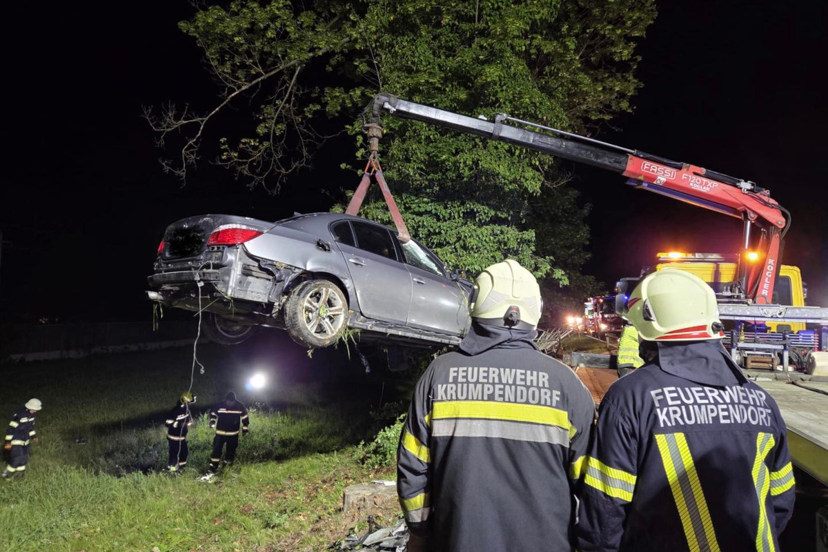 Ein Auto wird bei Nacht von einem Kran angehoben. Feuerwehrleute in Schutzkleidung beobachten die Szene. Die Umgebung ist dunkel und schlecht beleuchtet.