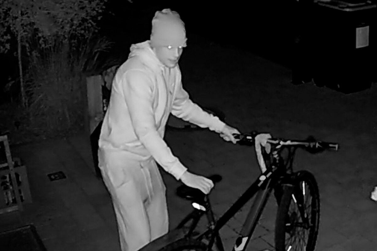 Eine Person in dunkler Kleidung schiebt ein Fahrrad auf einem Gehweg bei Nacht. Die Szene ist in Schwarzweiß gehalten.
