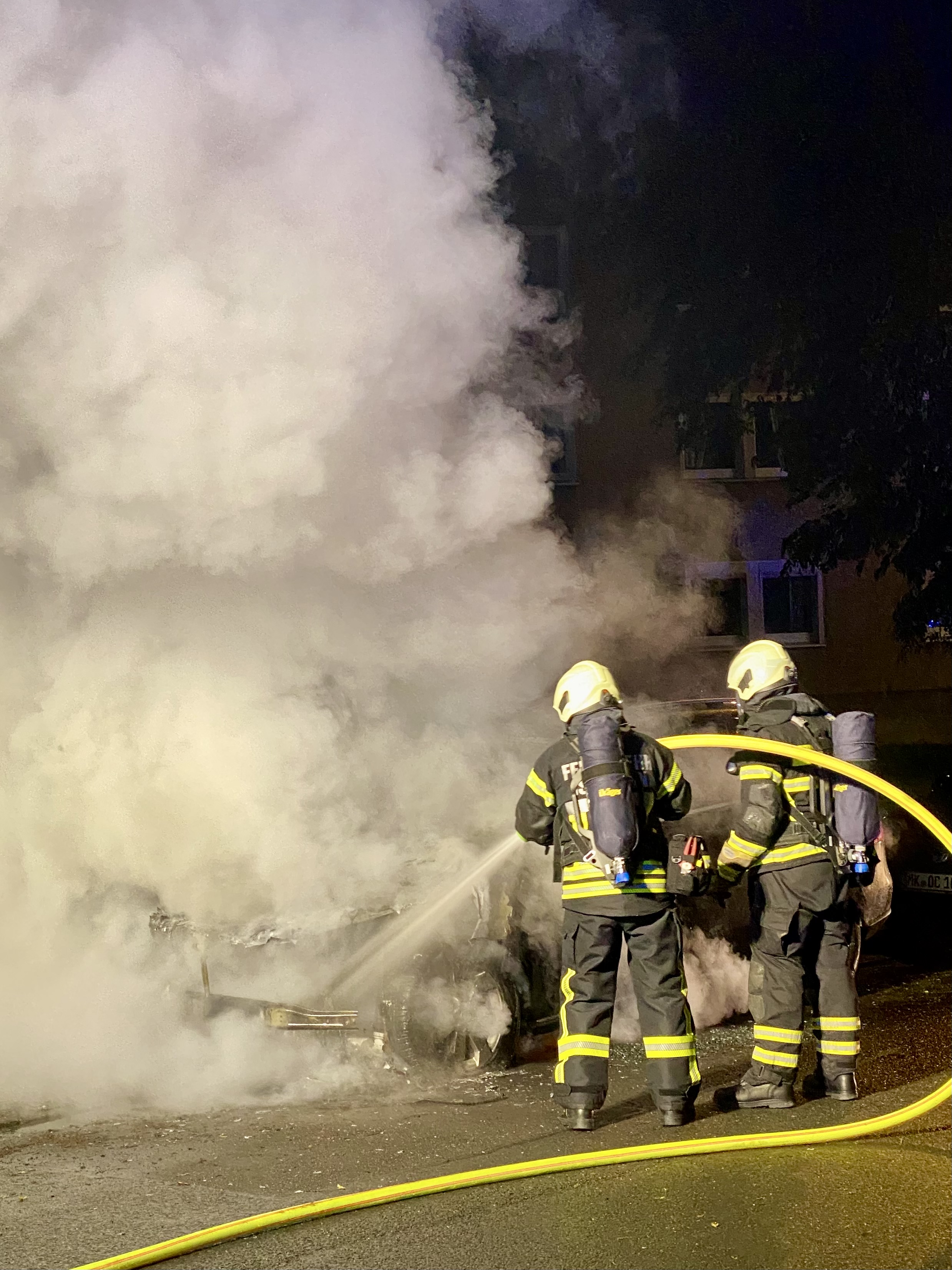Zwei Feuerwehrleute bekämpfen einen Autowrackbrand in einer rauchigen Umgebung. Das Fahrzeug ist stark beschädigt und von Rauch umgeben.