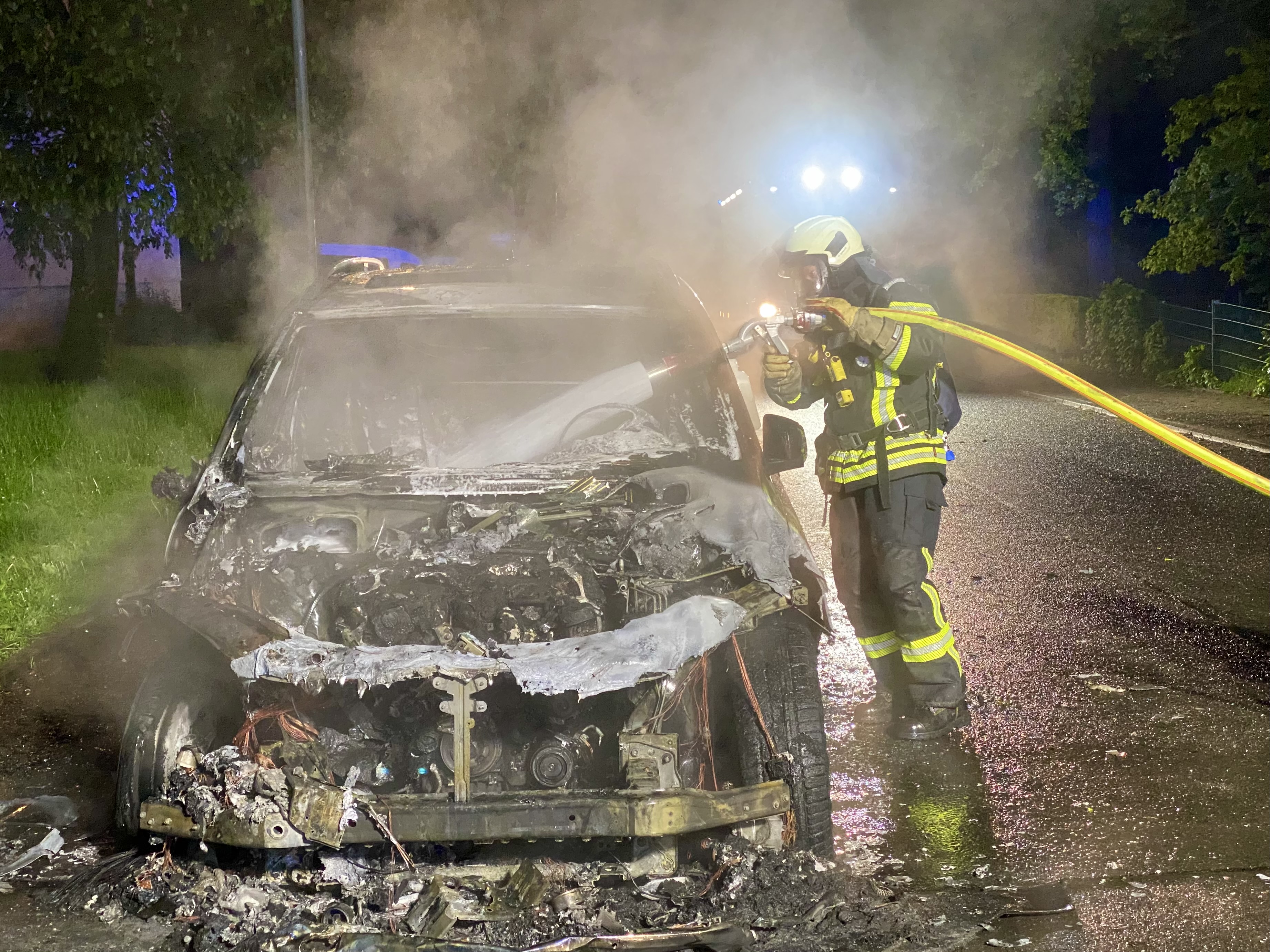 Ein Feuerwehrmann in voller Montur löscht mit einem Schlauch ein brennendes Auto bei Nacht auf einer nassen Straße.