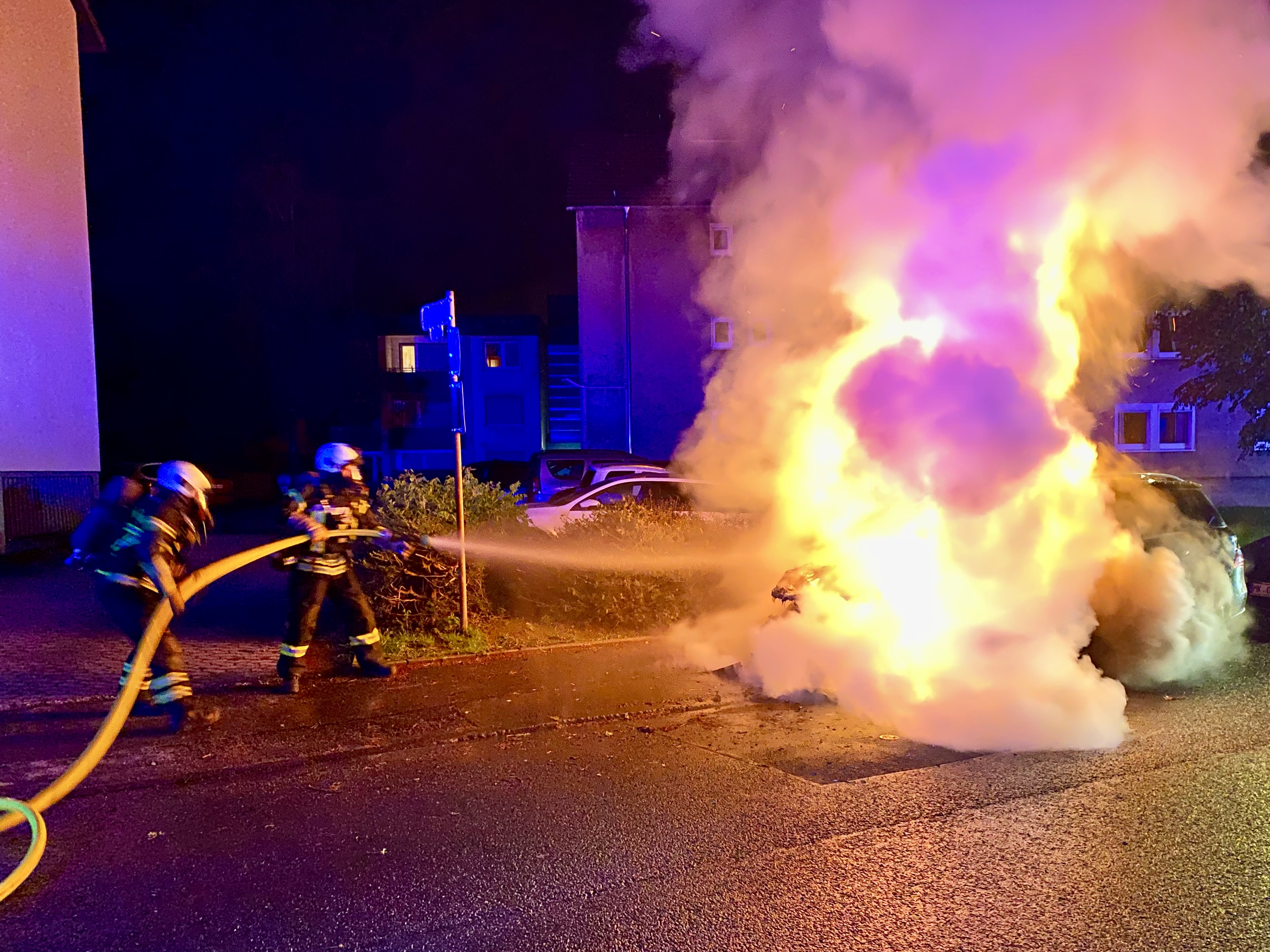 Zwei Feuerwehrleute in Uniform und Helmen sind dabei, ein brennendes Auto mit einem Wasserschlauch zu löschen. Das Auto steht am Straßenrand und ist von großen Flammen und Rauch umgeben.