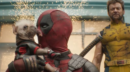 Im Vordergrund Deadpool in seinem anzug, der von einem kleinen, haarlosen Hund mit langer Zunge im Gesischt abgeschlabbet wird. Im Hintegrund Wolverine in gelbem Dress, der eine grimmige Fratze schneidet. Über allem fliegen kleine Funken.