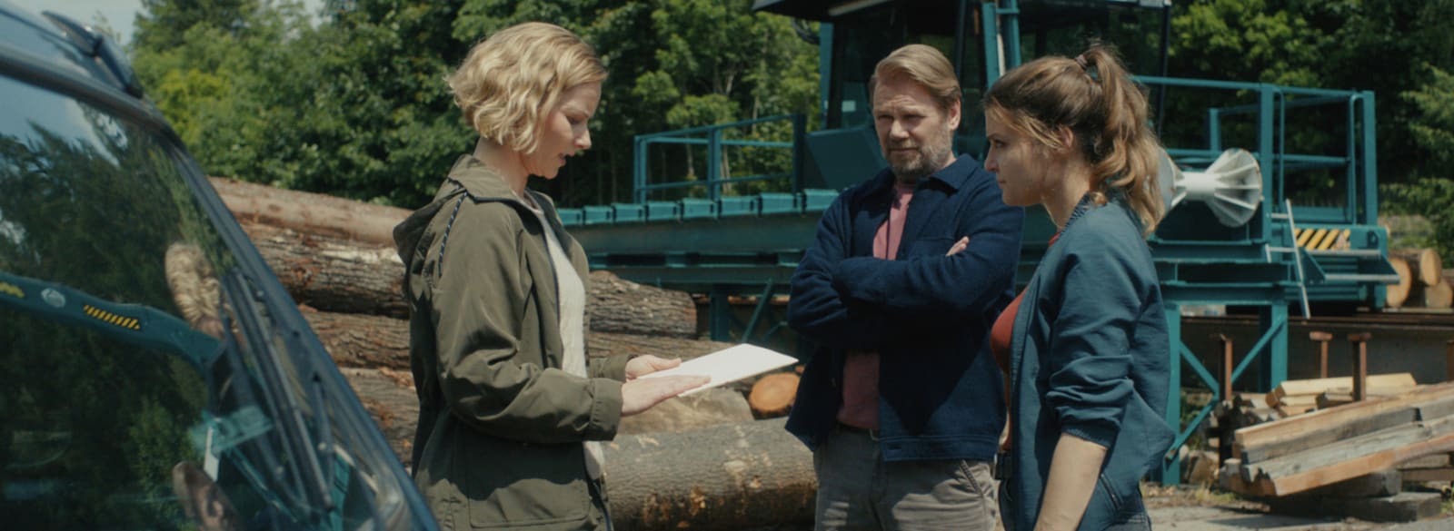 Auf einem Gelände mit schweren Holzmaschinen und aufgestapelten Stämmen bespricht Saskia Bergelt (Teresa Weißbach) mit Kommissar Winkler (Kai Scheve) und Kollegin Szabo (Lara Mandoki) wichtige Unterlagen.