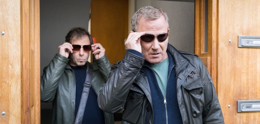 Ekki Talkötter (Oliver Korittke) und Overbeck (Roland Jankowsky) kommen gemeinsam aus einem Hauseingang. Beide tragen Lederjacken und setzen sich im Gehen gerade eine dunkle Sonnenbrille auf.