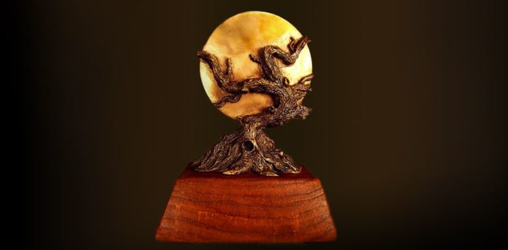 Statue des World Fantasy Award. Ein verschlungener dunkelbrauner Baum, der auf einem hellbraunen Holzsockel steht und eine goldene Scheibe umschlingt.