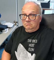 Dirk Van den Boom mit Brille an einem Schreibtisch sitzend. Trägt ein schwarzes Dune-T-Shirt mit einem Sandwurm darauf und dem Schriftzug "The Spice Must Flow"