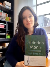 Paula Hauch mit der Neuausgabe von Heinrich Manns Roman »Der Untertan« vor einem Fenster und einem Bücherregal