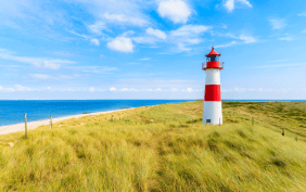 Foto vom Meer, davor Dünen und ein Leuchtturm bei schönem Wetter