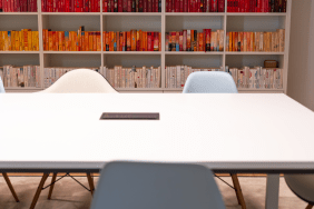 Ein großer weißer Besprechungstisch mit Stühlen im Vordergrund, dahinter eine Regalwand mit nach Farben sortierten Büchern in Rot, Orange und Weiß.