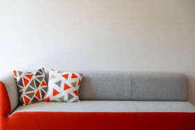 Ein moderner Sofa-Zweisitzer mit grauer Sitzfläche und knallorangefarbener Verkleidung, dekoriert mit zwei Kissen im geometrischen Muster vor einer schlichten weißen Wand.