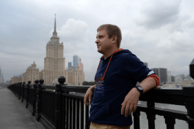 Der russische Autor Sergej Lebedew lehnt auf einer Brücke an einem Geländer, im Hintergrund ein großes, monumentales Gebäude aus Stein.