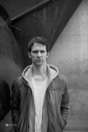 Der Autor Thomas von Steinaecker schaut in die Kamera. Er steht vor einem strukturierten Hintergrund aus Beton und trägt ein weißes T-Shirt unter einem offenen Kapuzenpullover mit Reißverschluss und einer Jacke.