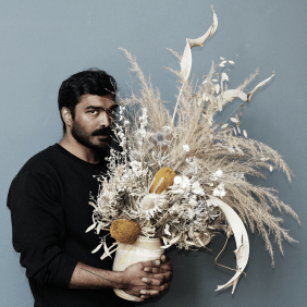 Varatharajah Senthuran steht schwarz bekleidet vor einer grauen Wand. In den Händen hält er eine große Vase, in welcher getrockneten Blumen stecken.