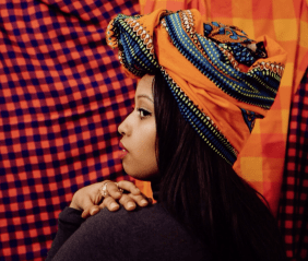 Die Autorin Warsan Shire schaut über ihre Schulter, ihre Hände liegen gefaltet auf ihrer Schulter, sie trägt einen bunten Kopfschmuck und schwarze Kleidung