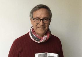 Hans Jürgen Balmes zeigt ein aufgeklapptes und schwarz-weiß-bebildertes Buch in die Kamera. Er trägt einen weinroten Pullover, Halstuch und eine Brille. Er lächelt leicht.
