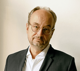 Autor und Journalist Nils Minkmar mit runder Brille, Dreitagebart und weißem Hemd und Anzugsakko vor einem hellen Hintergrund
