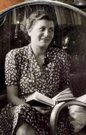 Schwarzweißfoto von Hedy. Sie lächelt und hält ein Buch im Schoß, während sie in einem Sessel sitzt. Blattgemustertes Kleid.