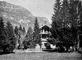 Schwarzweißfoto: Hotel Amalie in Garmisch-Partenkirchen. Holzhaus mit zwei Balkonen, umgeben von Tannen, vor Bergkulisse. Holzzaun im Vordergrund.