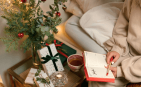 Frau sitzt im Anschnitt mit einem Pullover bekleidet auf einem Bett und hält ein Buch und einen Stift in der Hand. Die Szenerie ist weihnachtlich dekoriert