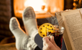 Eine Person trinkt einen Glühwein, hat ein Buch in der Hand. Im Hintergrund sieht man die Füße mit Kuschelsocken und einen Kamin mit Feuer
