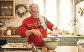 Ein älterer Mann mit Weihnachtspullover steht in der weihnachtlich dekorierten Küche und bäckt Plätzchen