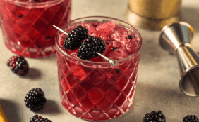Bramble Cocktail mit Brombeeren garniert, im Hintergrund Brombeeren und Cocktail-Werkzeug