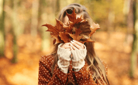Frau hält sich im herbstlichen Wald große Herbstblätter vor das Gesicht